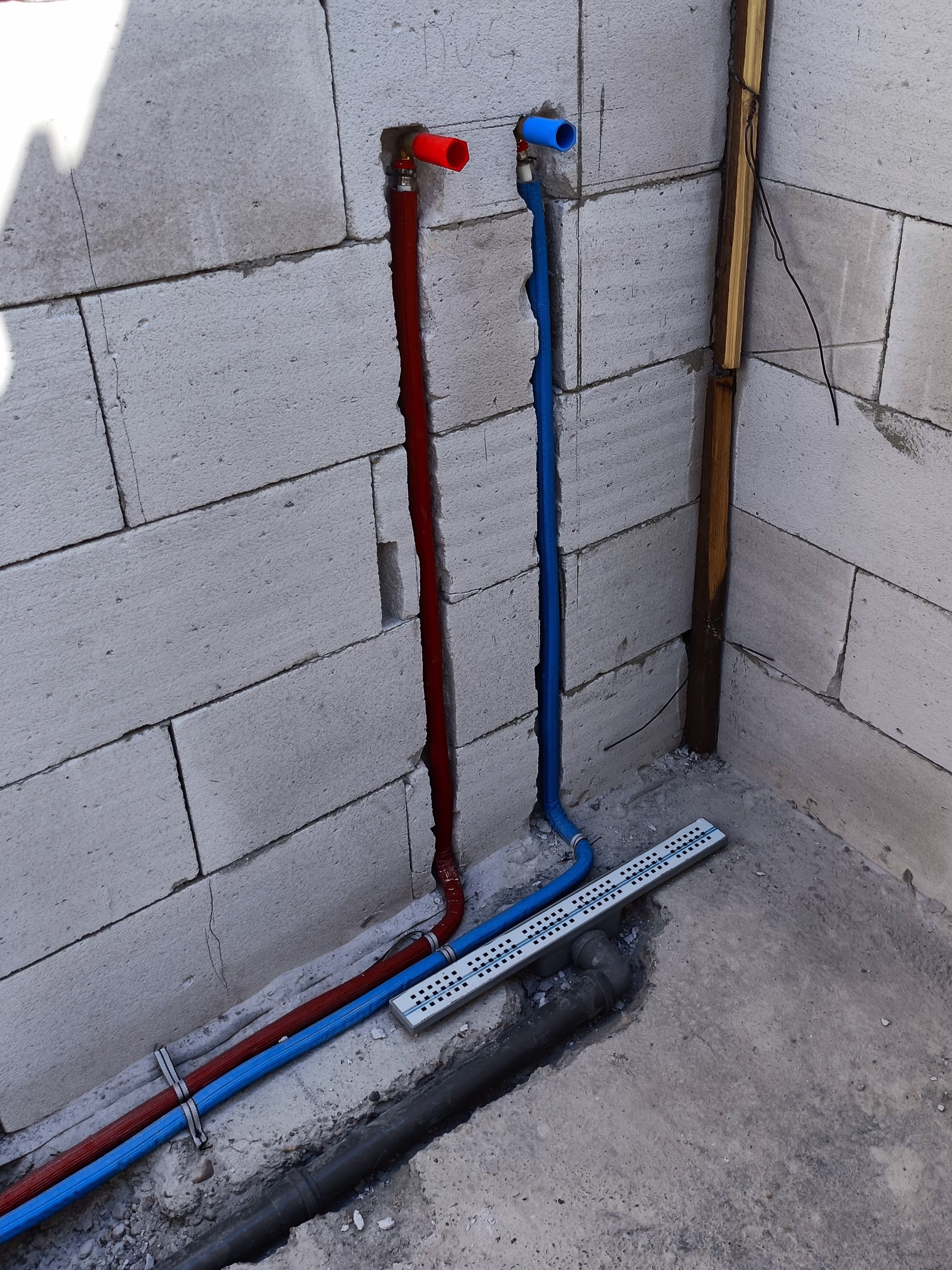 Exposed red and blue water pipes embedded in a concrete wall, positioned near a floor drain system under construction.