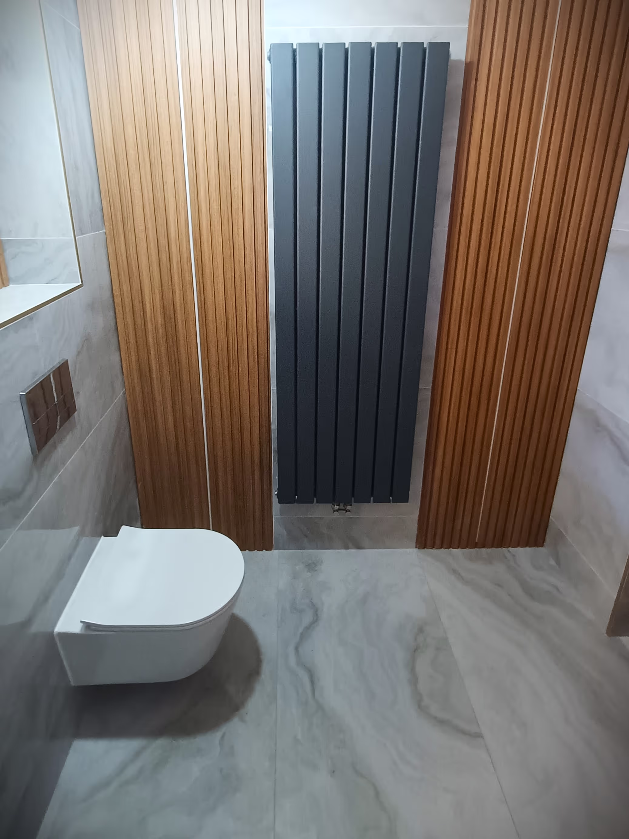 A modern bathroom with a floating toilet, marble walls, vertical wooden paneling, and a sleek black radiator mounted on the wall.