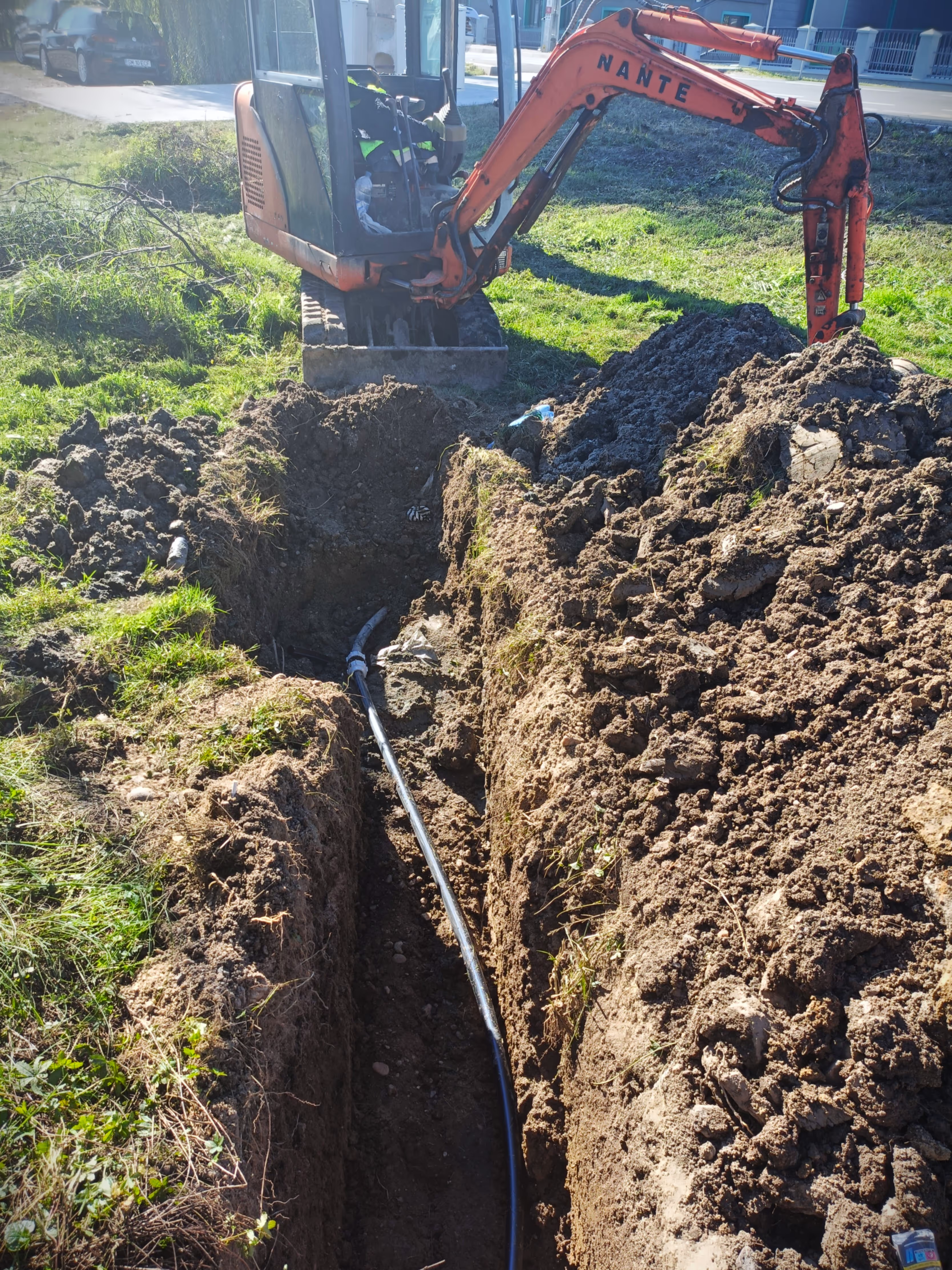 A small excavator digging a trench for underground utilities, with a black water pipe already partially laid in the soil.