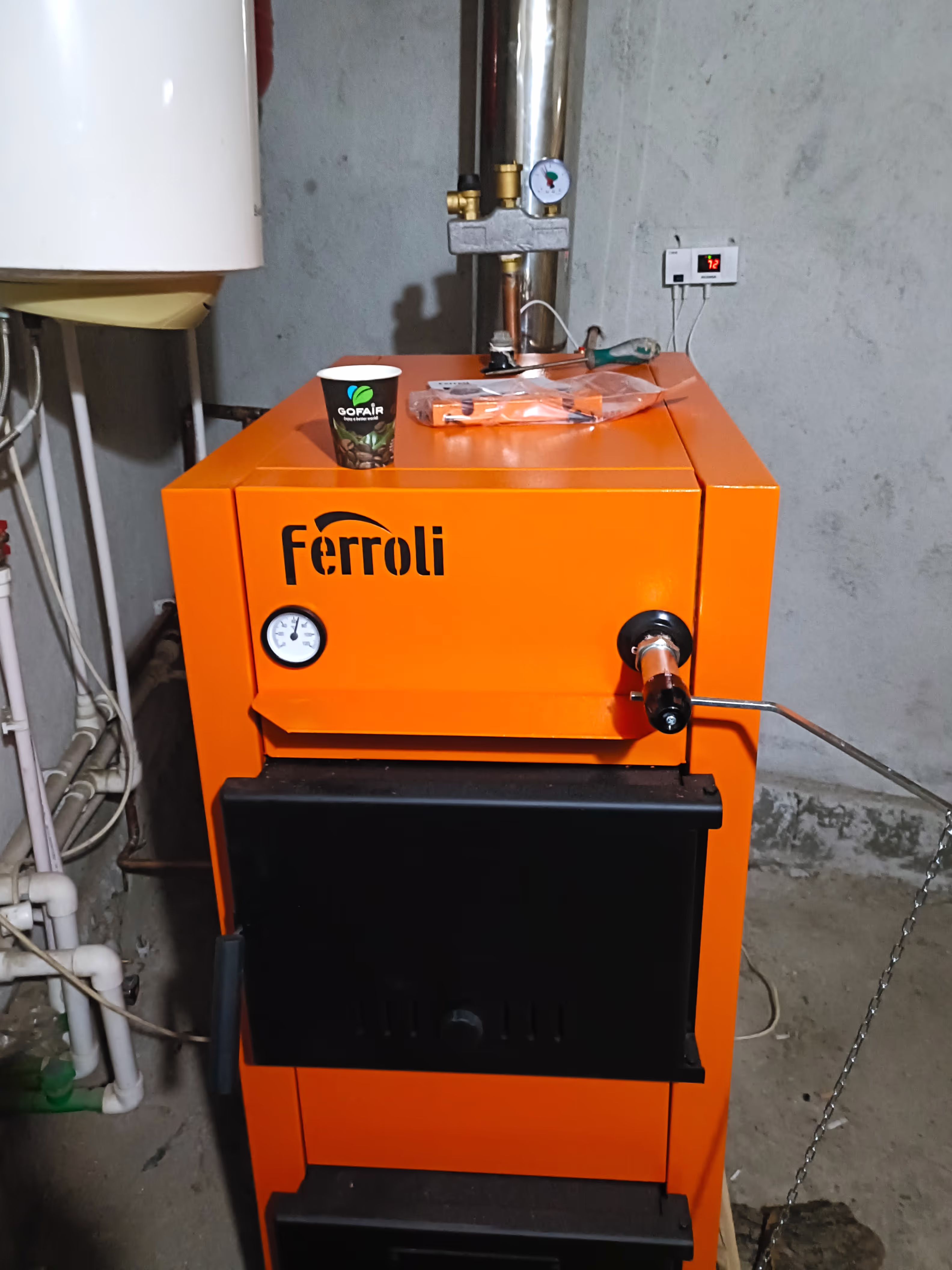 A Ferroli-brand solid fuel boiler with an orange metal body, black doors, and a pressure gauge, installed in a utility room with various pipes and a white water tank.