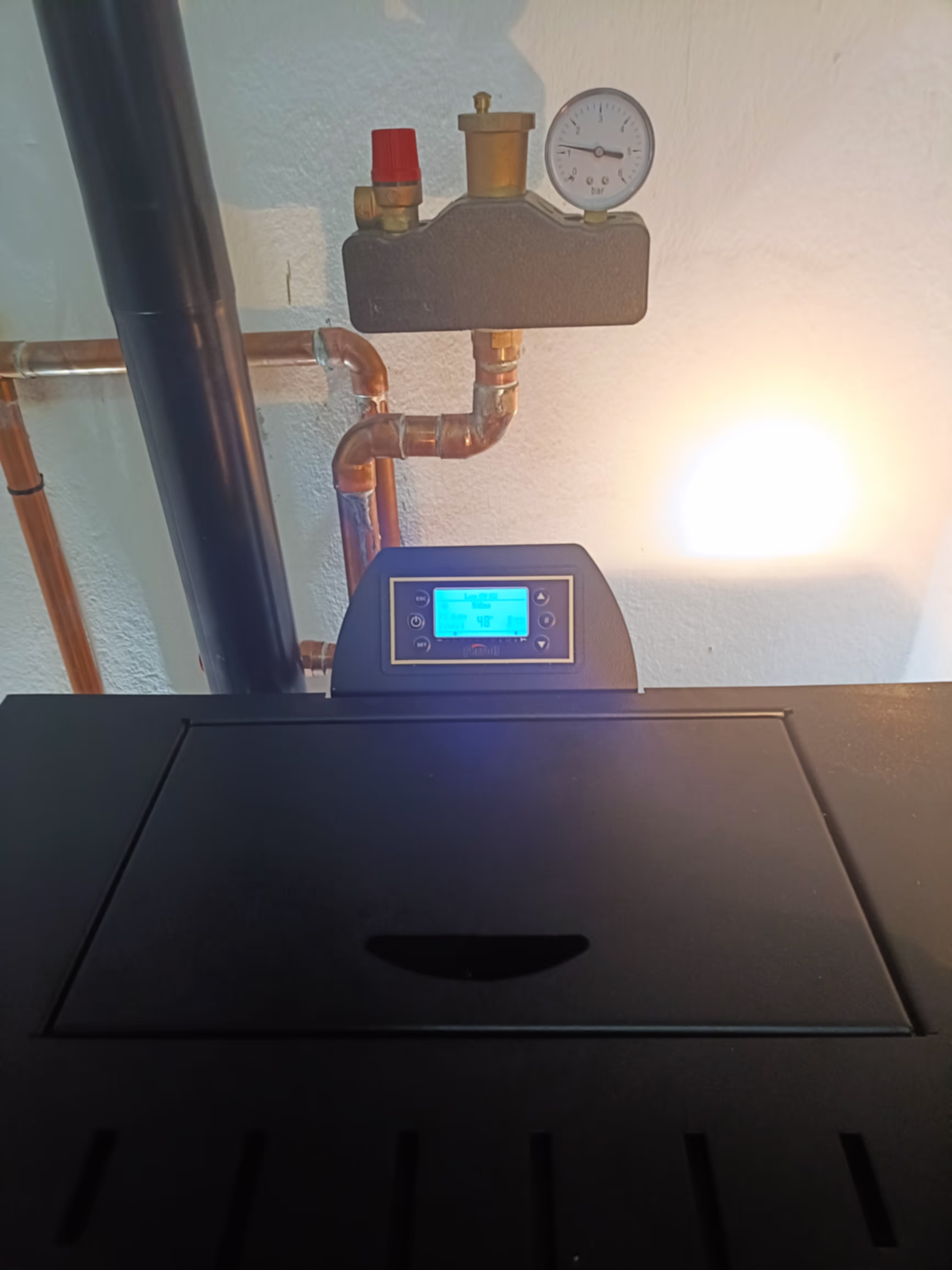 A digital control panel on top of the Ferroli heating stove, with connected pipes and a pressure gauge.
