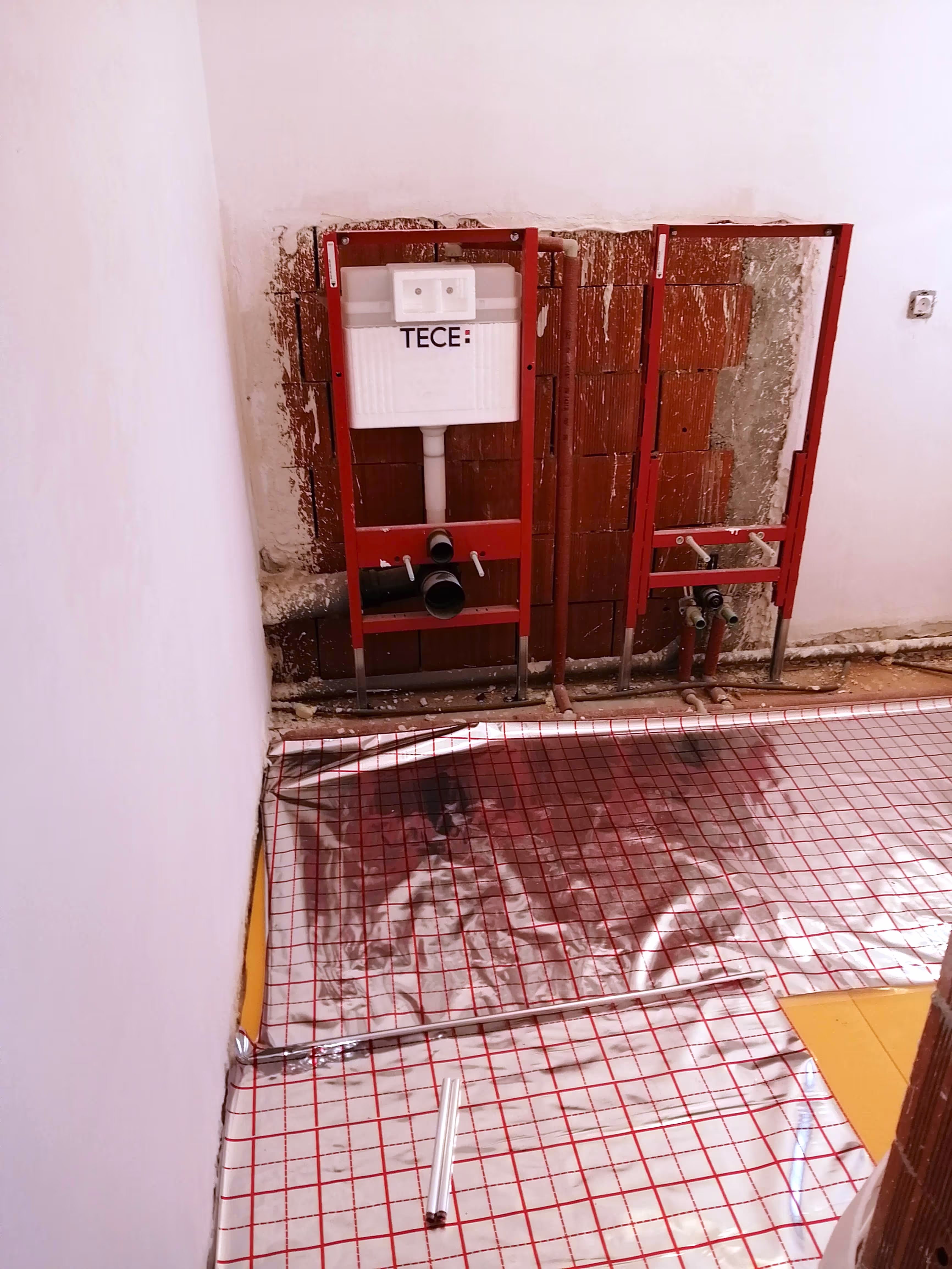 A bathroom installation in progress, showing two red TECE toilet frames mounted in a brick wall, with underfloor heating insulation laid out.