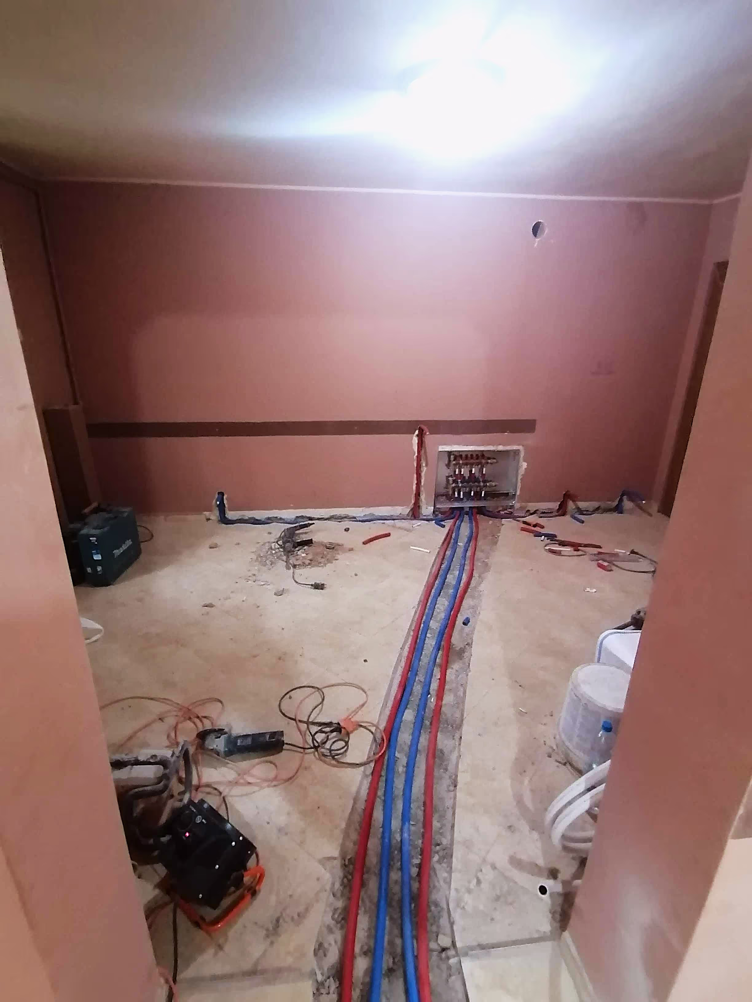 A partially constructed room with heating pipes in red and blue embedded in the floor, leading to a distribution panel in the wall.