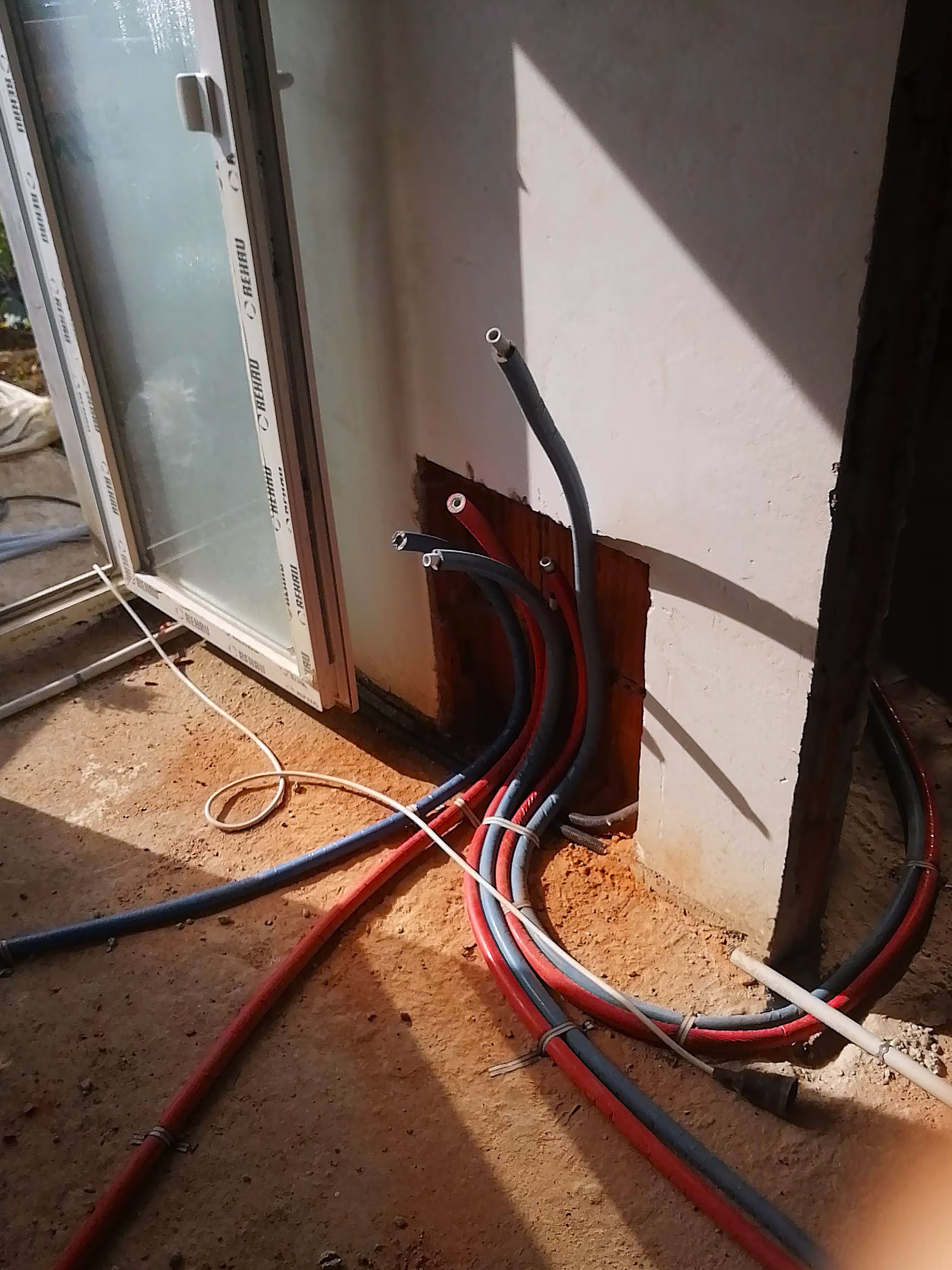 Exposed heating pipes in red and blue emerging from a wall near a glass door, showing early-stage plumbing installation.