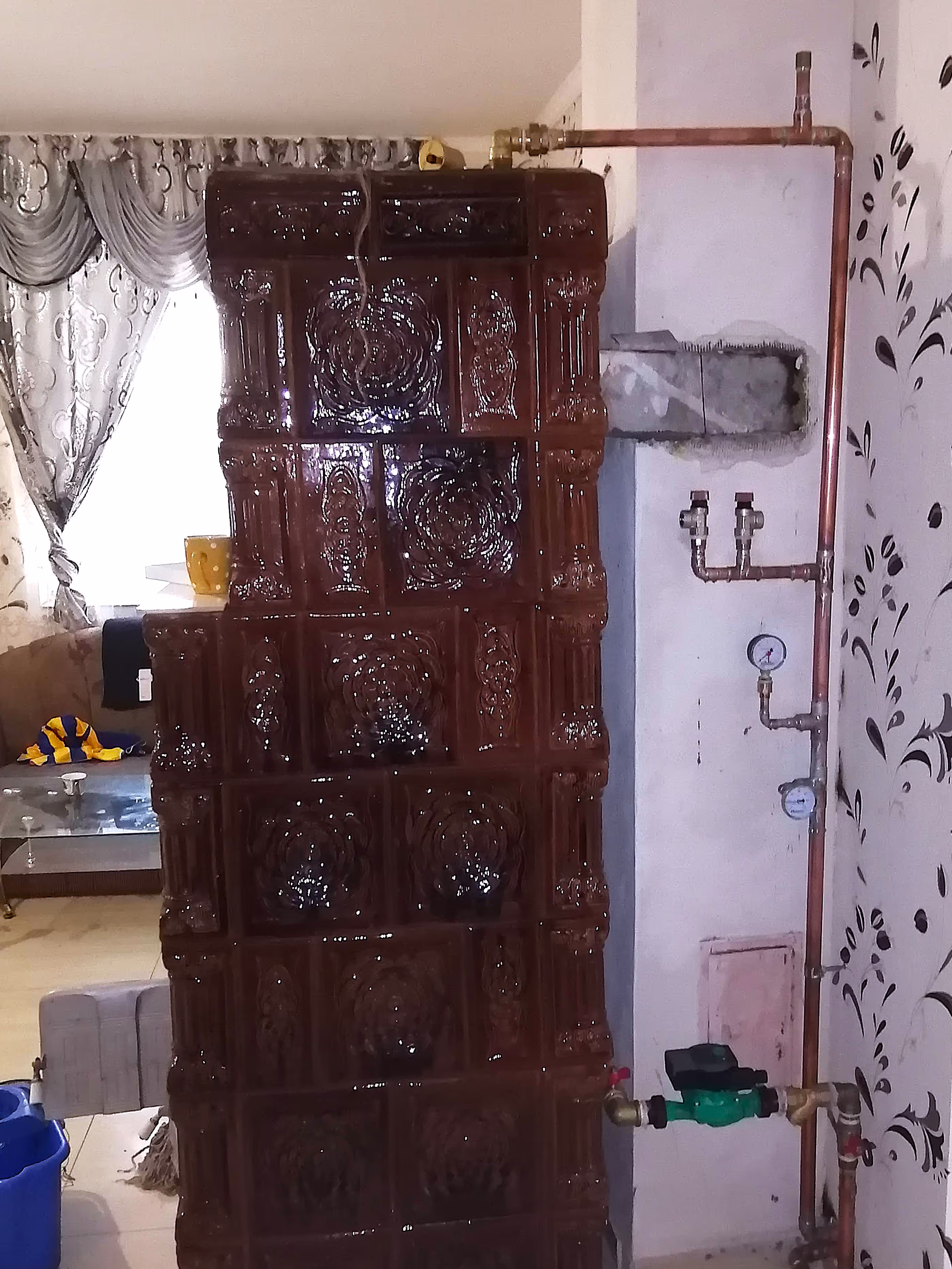A traditional tiled ceramic stove with a copper pipe heating system connected to it, installed in a living space with a curtain and furniture.