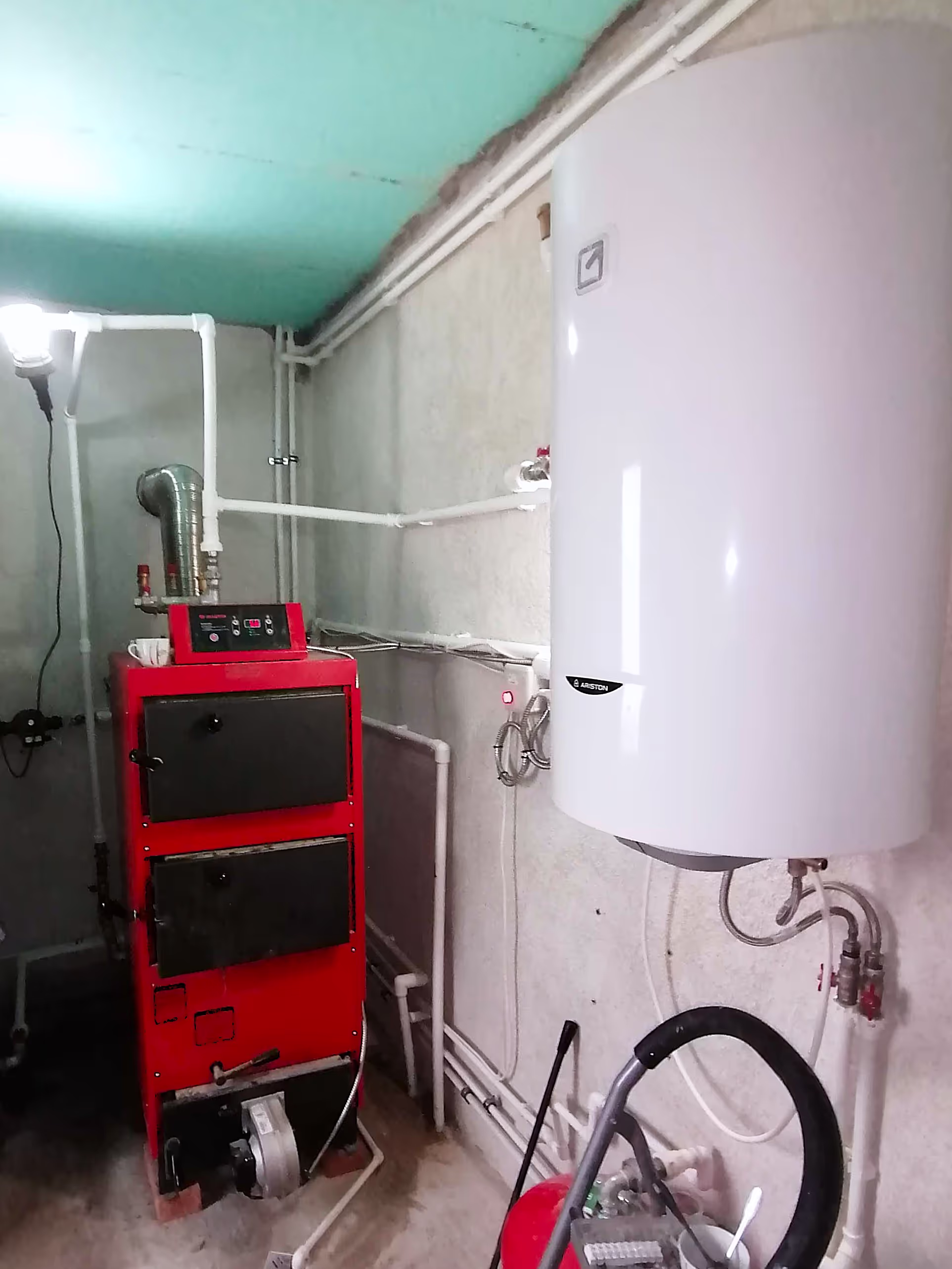 A red and black solid fuel boiler installed in a utility room with white PVC pipes running along the wall and ceiling, next to a white water heater.