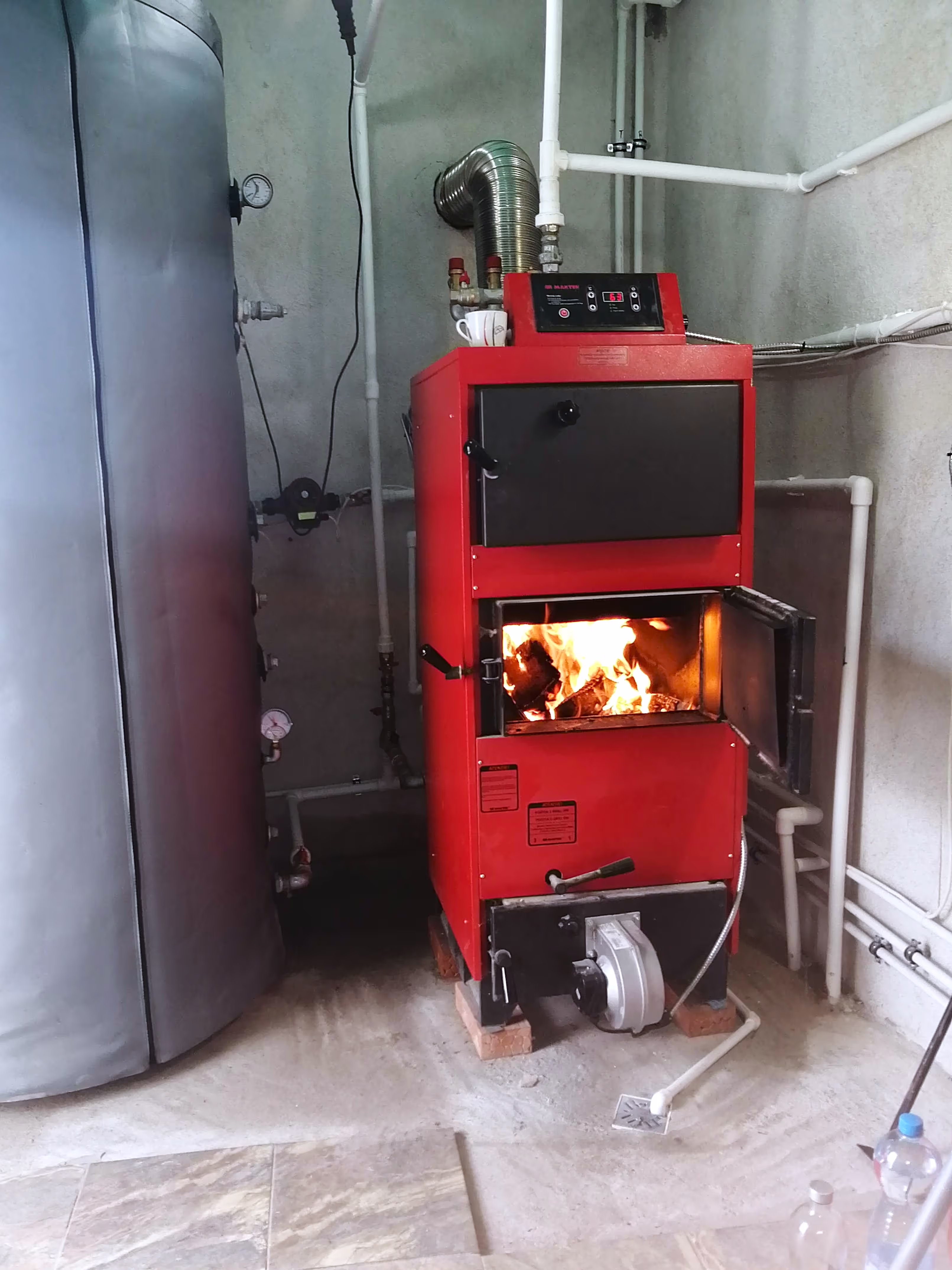 A red solid fuel boiler with its front door open, showing a fire burning inside. A large insulated storage tank is visible beside it.