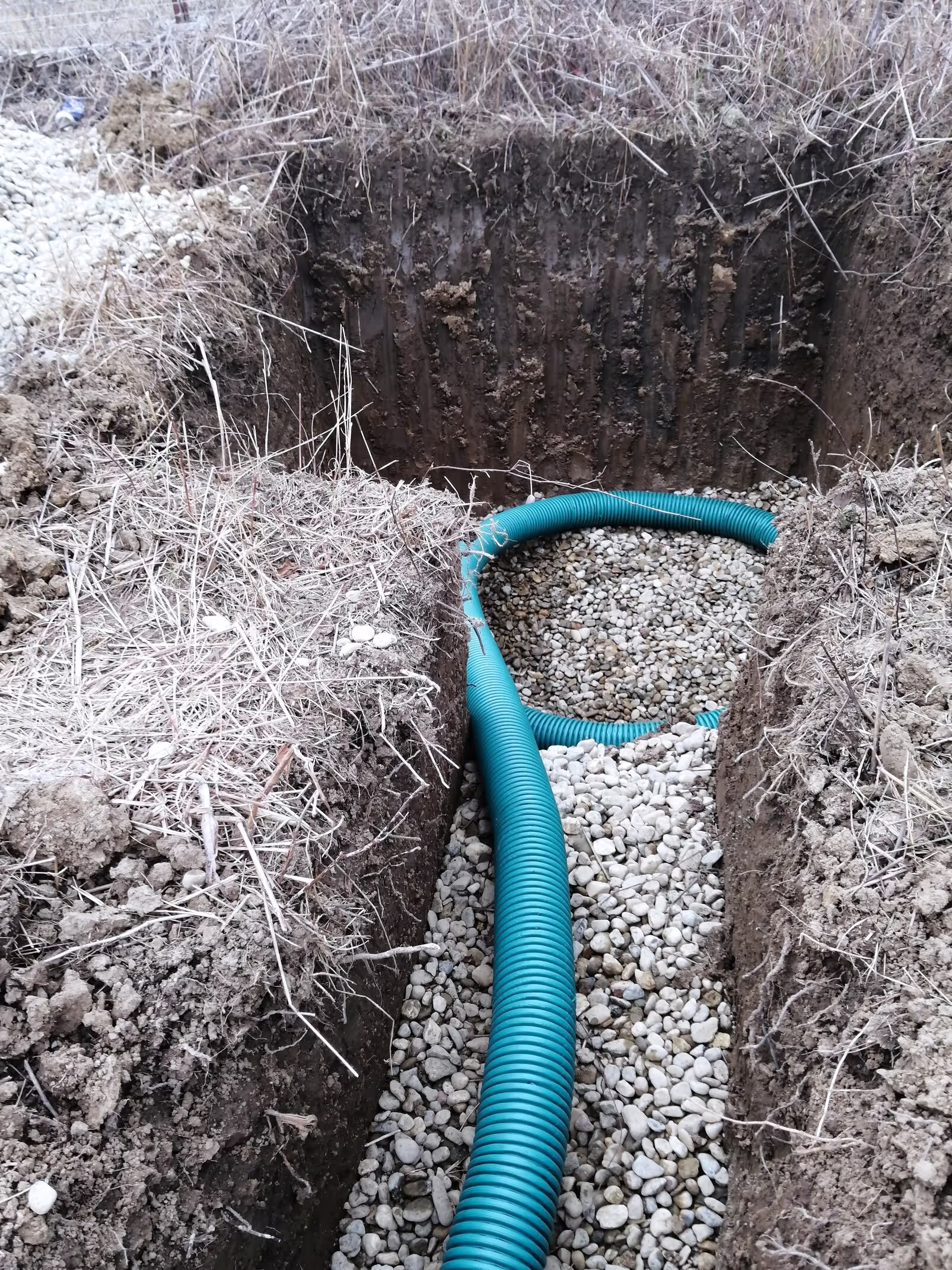 A dug-out trench filled with gravel and a flexible blue corrugated drainage pipe partially buried in the stones.