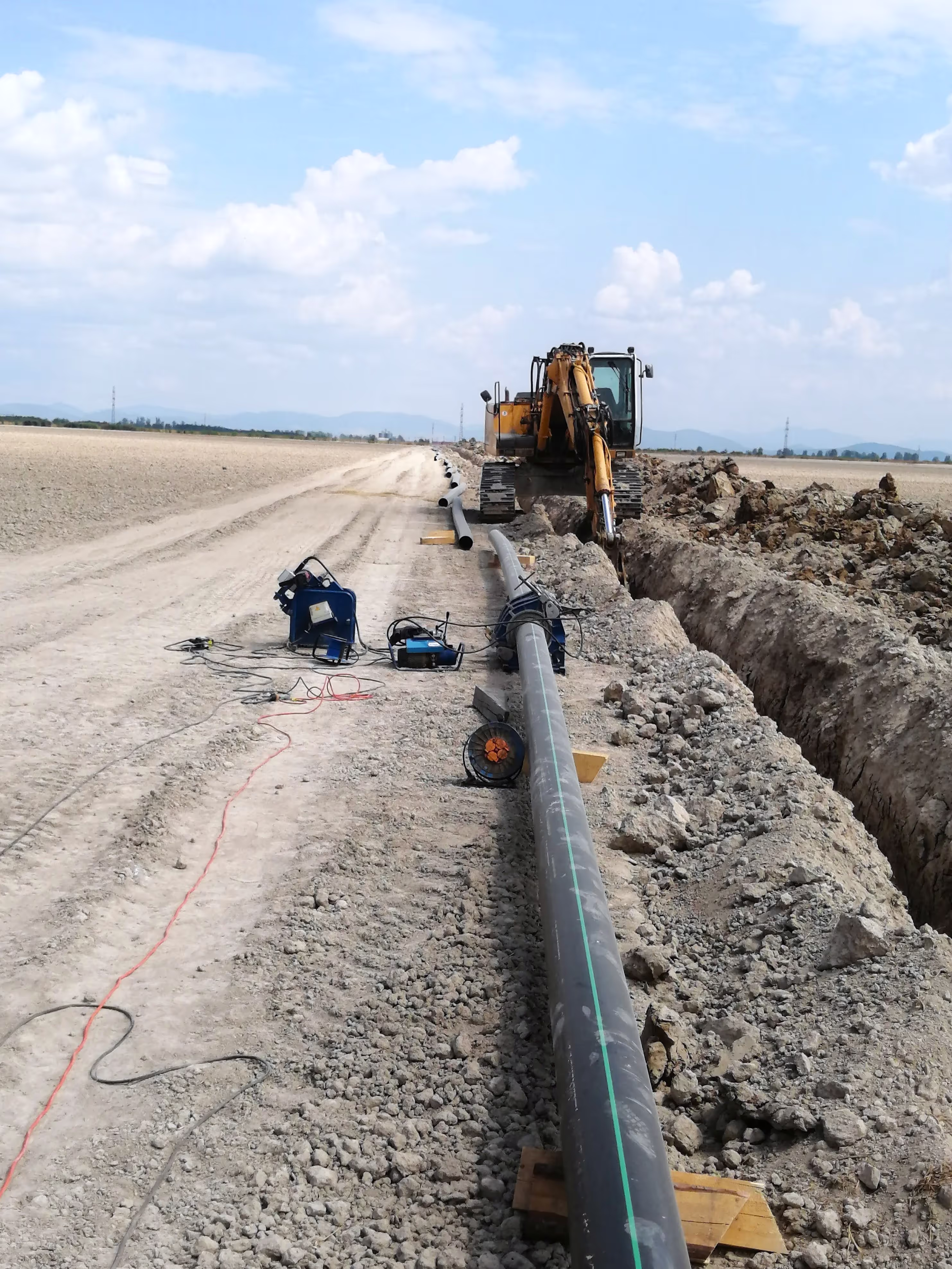 A backhoe excavator working on a trench for pipeline installation, with welding equipment and large pipes aligned along the site.