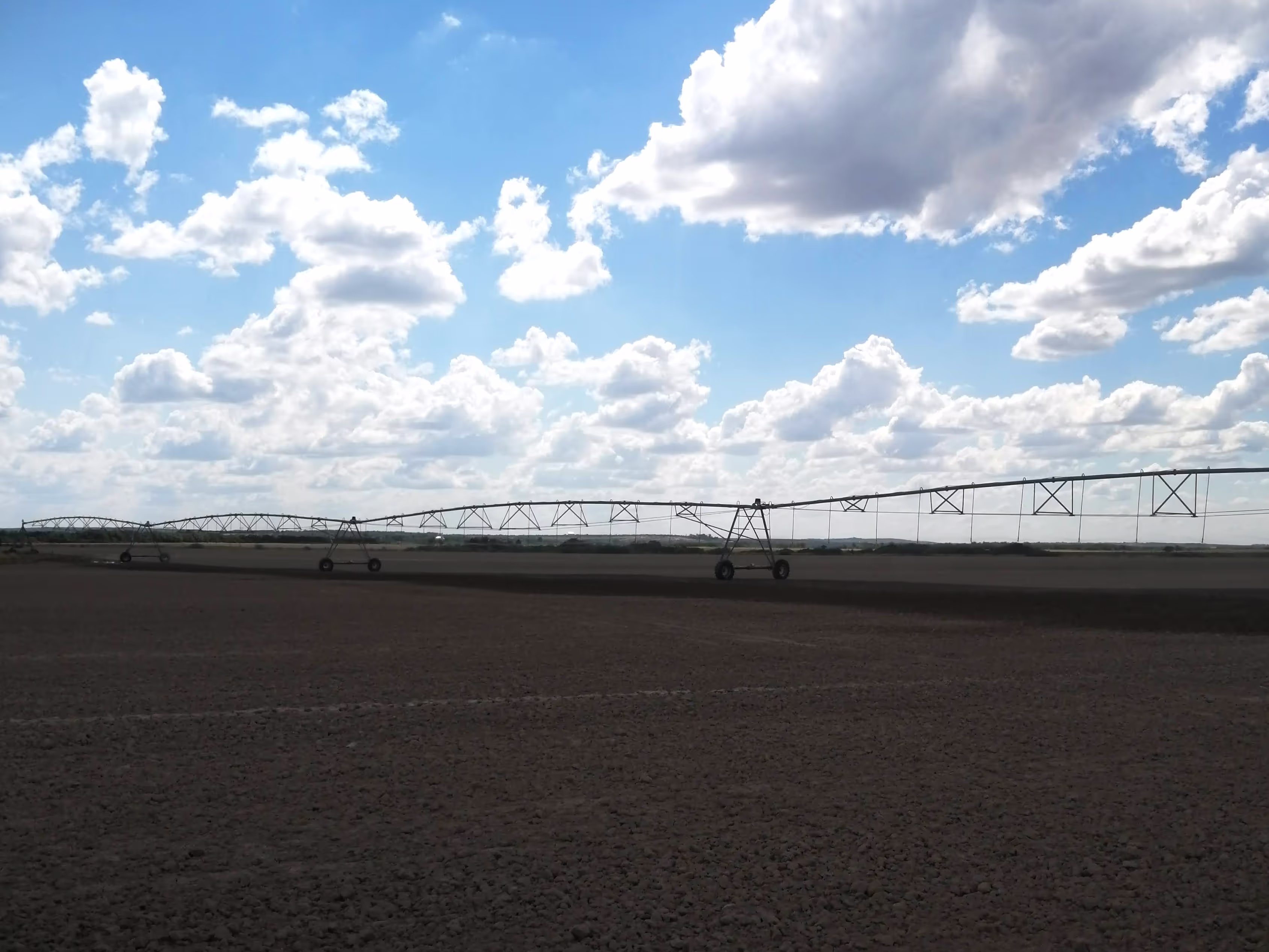 A vast field with a long center pivot irrigation system extending under a bright sky with scattered clouds.
