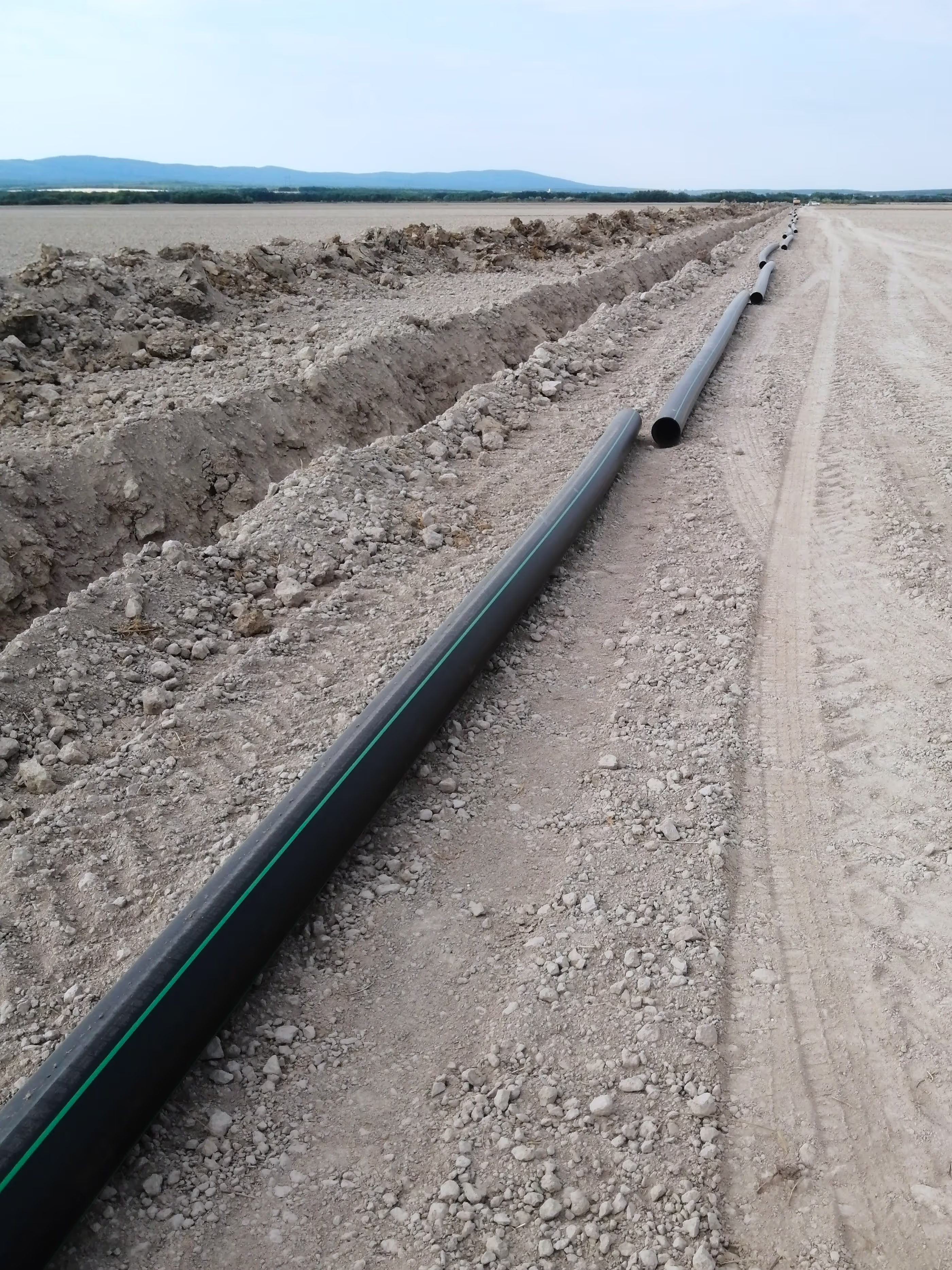A long black pipeline laid on a dry construction site, stretching into the distance with visible trench work.