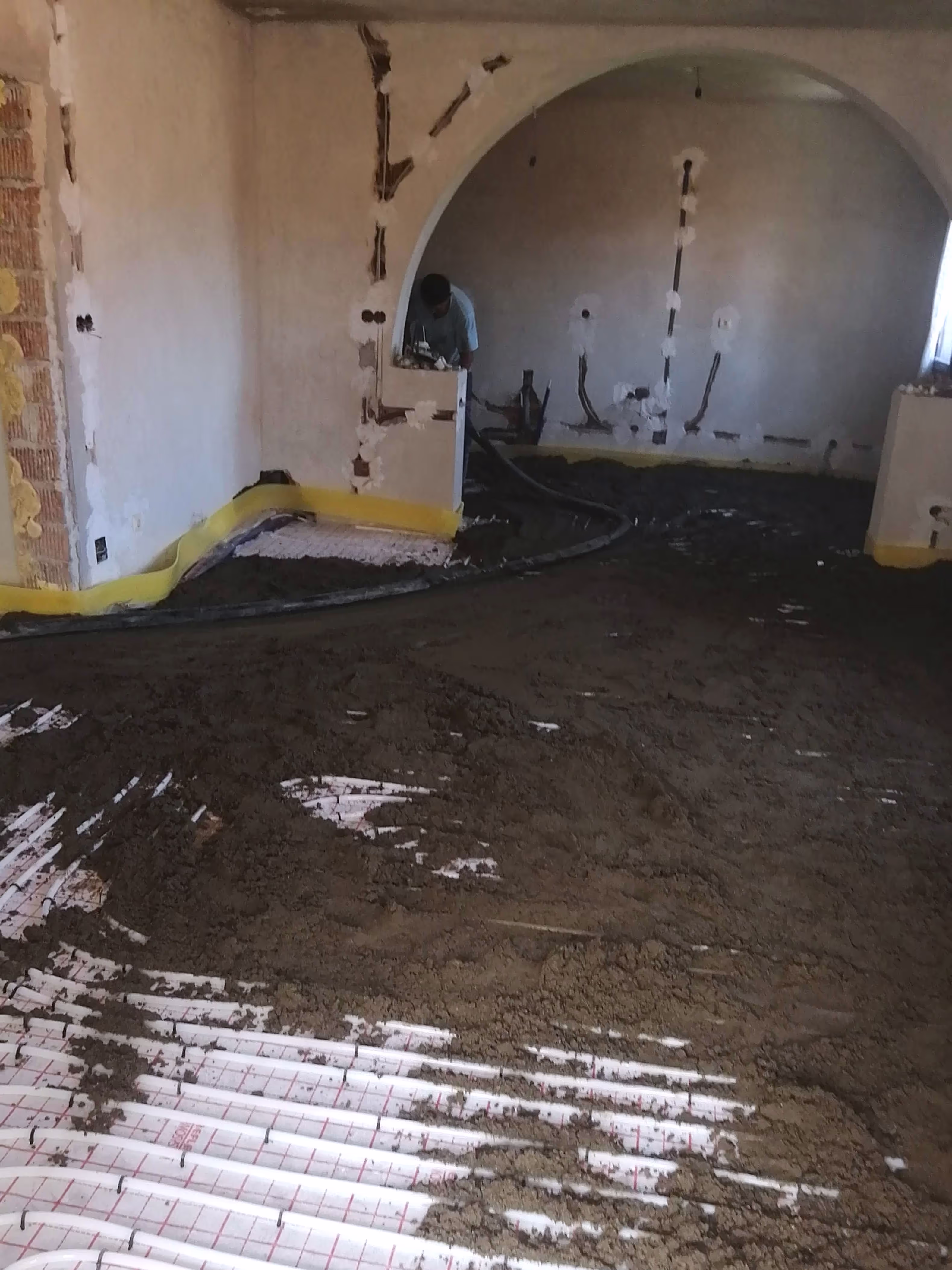 A room with underfloor heating with a worker pouring and spreading a concrete screed over the pipes.