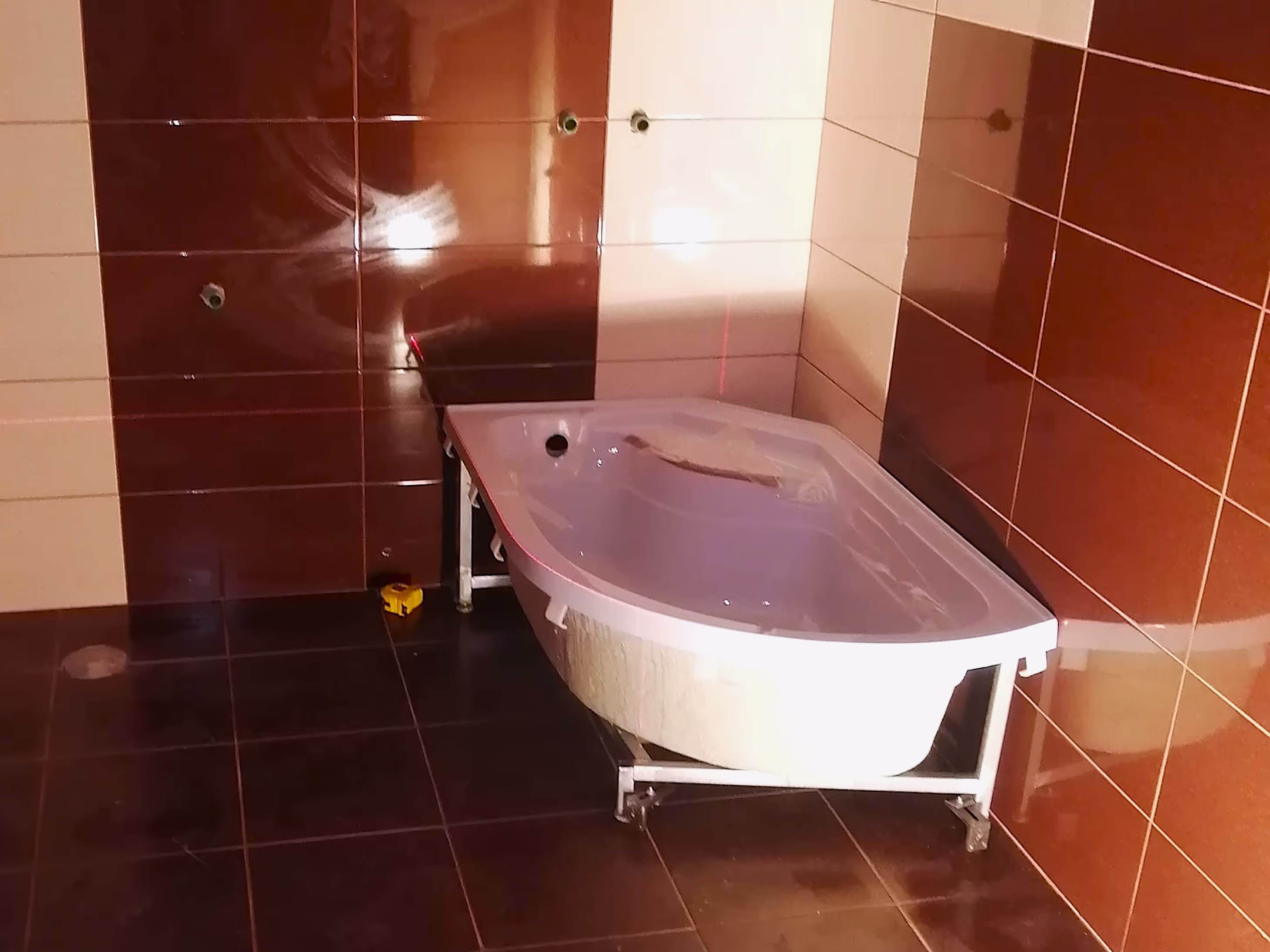 A bathroom under construction with a partially installed corner bathtub, brown and beige tiled walls, and laser level markers for alignment.